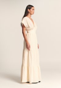 Ærmeløs maxikjole i cremefarve med dyb V-hals, tekstureret stof og lagdelt nederdel. Har en tætsiddende talje og en flydende kant.