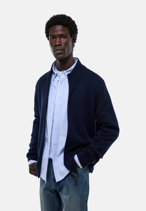 Un pull zippé bleu marine avec des poignets et un col côtelés, porté par-dessus une chemise à boutons bleu clair. Un jean en denim complète la tenue.