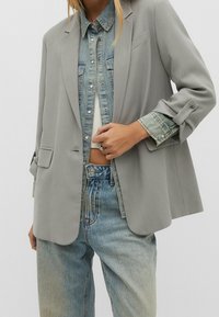 Femme portant un blazer gris clair par-dessus une veste en jean et un jean, montrant son ventre et une bague argentée en forme de cœur à la main.