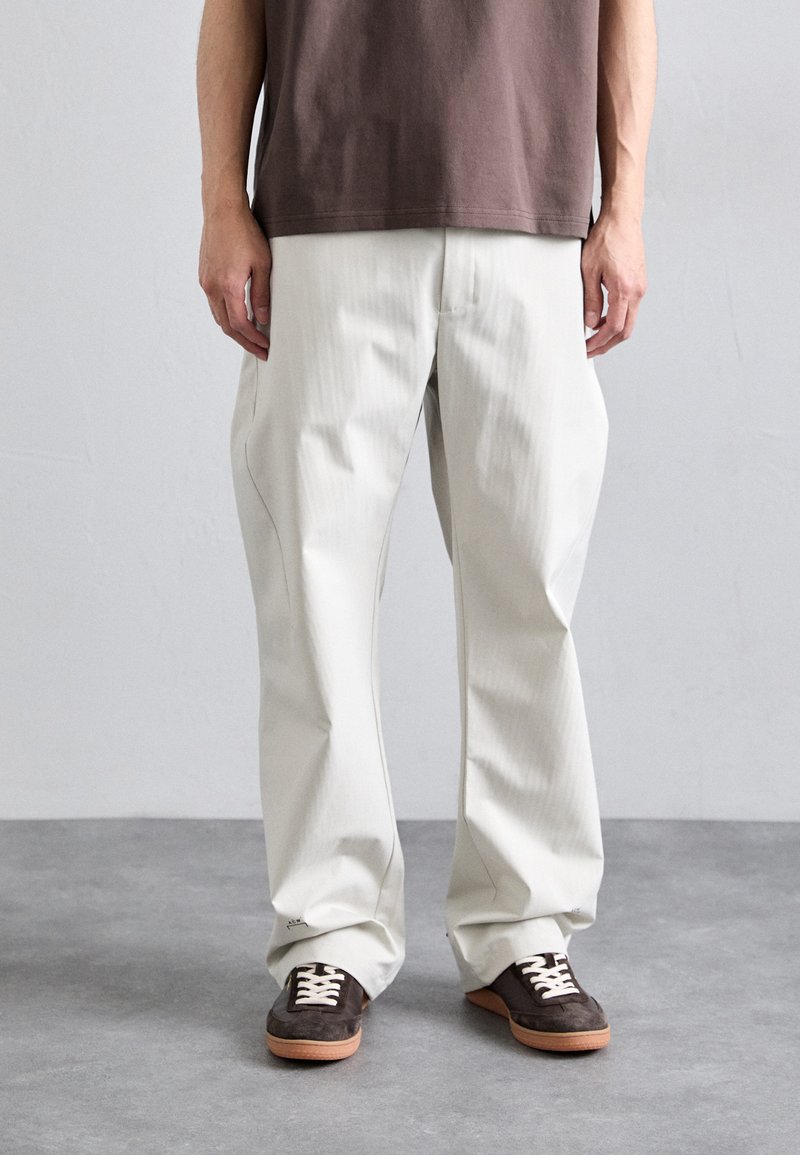 Weiße, plissierte Hose mit lockerer Passform, aus glattem Stoff hergestellt. Kombiniert mit braunen Sneakers mit weißen Schnürsenkeln und einer hellbraunen Sohle.