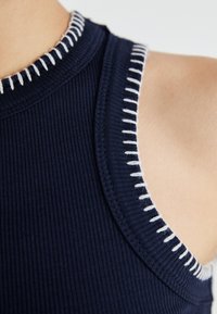 Camisola de alças de cor azul-marinho com costura branca contrastante ao longo do decote redondo e das cavas. Close-up da textura e dos detalhes do tecido.