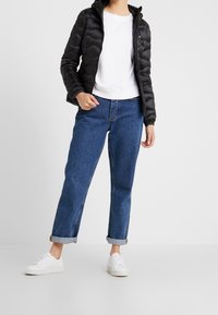 Schwarze Steppjacke mit einem gesteppten Design, weißes T-Shirt mit Rundhalsausschnitt, blaue cropped Jeans und weiße Sneaker mit Schnürsenkeln.