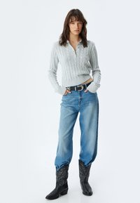 Pullover grigio chiaro a maglia con colletto, abbinato a jeans blu in denim larghi e stivali da cowboy neri in pelle con ricami.