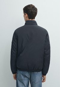 Czarna pikowana kurtka z wysokim kołnierzem, elastycznymi mankietami i dopasowanym dołem; gładka faktura i minimalistyczny design. Noszona z jasnoniebieskimi dżinsami.