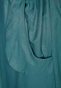 Teal fabric with a smooth texture, featuring a large, rounded pocket integrated into the side, subtle sheen, and minimalistic design.