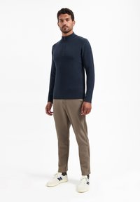 Marineblauwe ribbeltrui met hoge kraag en rits, gecombineerd met beige broeken en witte sneakers, met blauwe accenten en een gladde textuur.