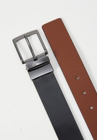 Vendbar belte med svart skinn på den ene siden og teksturert brunt skinn på den andre. Har en firkantet, matt svart metallspenne.