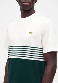 Personne à la peau foncée portant un t-shirt Lacoste rayé blanc et vert foncé avec un petit logo crocodile vert sur la poitrine.
