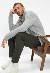 Grijze kabelgebreide cardigan, lichtgrijs t-shirt met ronde hals, olijfgroene broek, witte sneakers, zittend op een houten en leren stoel.