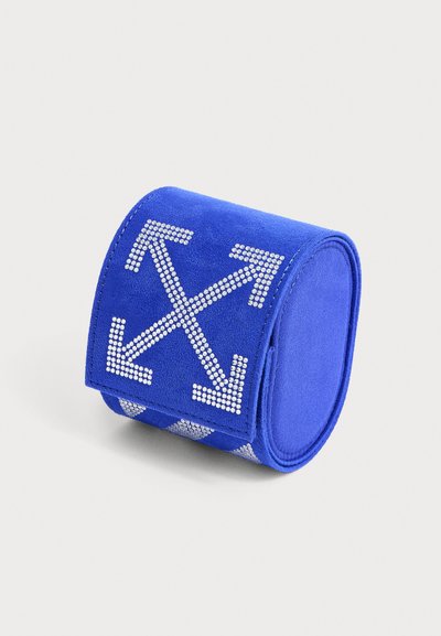 Bracelet manchette en daim bleu avec des flèches en strass blancs croisées en forme de X sur le dessus, présenté sur un fond clair uni.