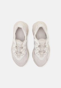 Sapatilhas de desporto em bege claro, com parte superior em malha e sintético, atacadores texturizados e sola robusta. Marcação apresentada na língua e na palmilha.