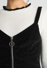 Svart kordflöjlsklänning med V-ringning och justerbara band, med en framdragkedja med ett runt draghandtag. Vit ribbad långärmad topp under.