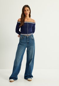 Marinblus med avskuren axel och vita ränder, med dragkedja, ihop med högmidjade blå vida jeans och beige sneakers, på en neutral bakgrund.