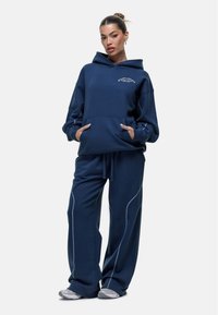 Marineblauwe fleece hoodie met een grote zak aan de voorkant en witte stikseldetails, gecombineerd met bijpassende losse marineblauwe sweatpants en grijze sneakers.