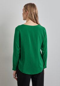 Blusa verde a maniche lunghe realizzata in un tessuto liscio, con scollatura rotonda e orlo leggermente curvo, indossata con pantaloni neri.