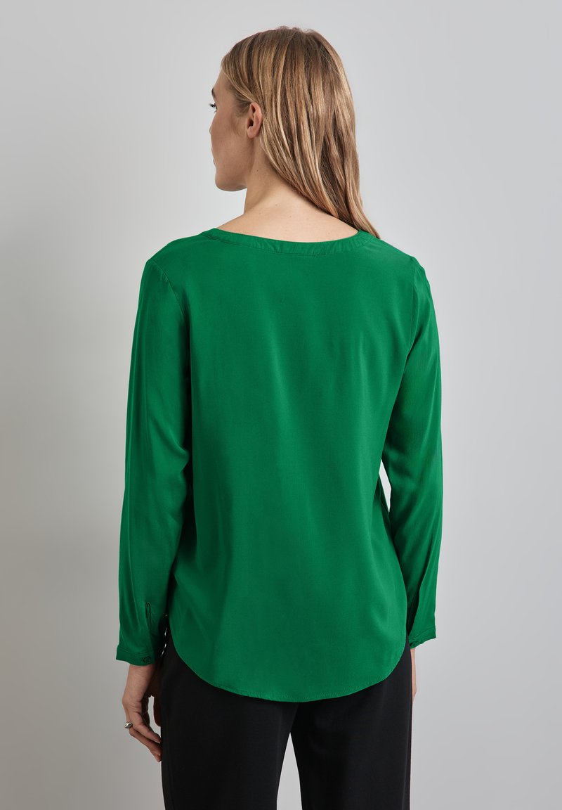 Blusa verde a maniche lunghe realizzata in un tessuto liscio, con scollatura rotonda e orlo leggermente curvo, indossata con pantaloni neri.