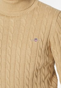 Beige Strickpullover mit Rollkragen, der gerippte Bündchen und einen Kragen hat. Kleines besticktes Wappen-Detail auf der linken Brust. Weicher Griff.