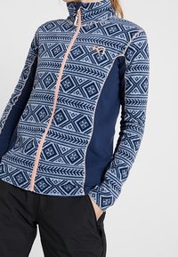 Granatowa i wzorzysta kurtka polarowa z wysokim kołnierzem, pełnym zamkiem błyskawicznym i kontrastowymi wykończeniami. Posiada geometryczne wzory w odcieniach niebieskiego i różowego.