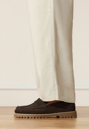 Beige pants paired with dark brown suede loafers featuring thick tan rubber soles and stitching details, standing on a wooden floor.