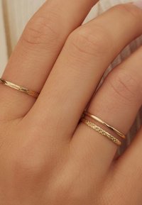 Trois délicates bagues en or sur une main à la peau claire. Les bagues varient en texture : l'une a une finition lisse, une autre est tordue, et l'autre est tressée.