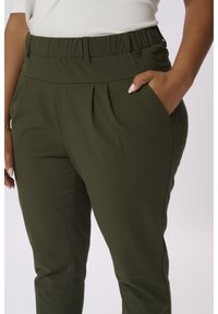 Personne portant un pantalon vert olive avec des plis et une taille élastique, la main placée dans la poche droite, sur un fond uni.