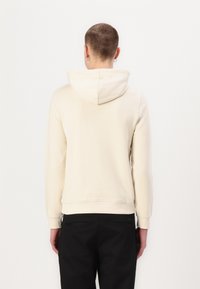 Sudadera beige hecha de una tela suave, con capucha y puños acanalados. La parte posterior es lisa con un detalle mínimo. Lleva puesta sobre pantalones negros.