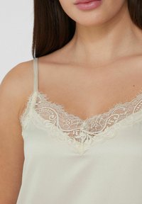 Camiseta de seda color beige claro con tirantes ajustables, que presenta un delicado encaje alrededor del escote. Textura suave y diseño en V.
