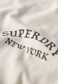 Camiseta de algodón blanca con texto negro que dice "superdry NEW YORK" en una tipografía audaz y de estilo vintage. Textura suave y lisa.