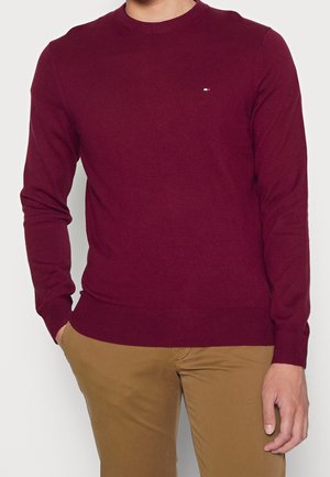 Man die een nauwsluitende bordeauxrode trui met lange mouwen en beige broek draagt, staand met één hand in de zak tegen een effen achtergrond.