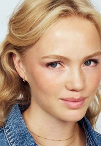 Ung kvinna med blont hår, lätt smink, iklädd små guldörhängen, lager av fina guldhalsband och en blå denimjacka.