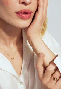 Närbild av en kvinna med ljus hy, som bär en vit skjorta och guldsmycken, försiktigt rör vid sin kind och handled med en hand.