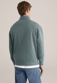 Svetlo zeleni pulover z visokim ovratnikom in dolgimi rokavi. Ima sproščen kroj, rebraste manšete in gladko teksturo. Nošen čez belo srajco.