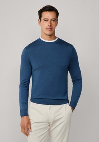 Blauer Langarm-Pullover mit Rundhalsausschnitt und strukturiertem Finish, kombiniert mit einem kontrastierenden weißen Kragen, getragen zu hellen Hosen.