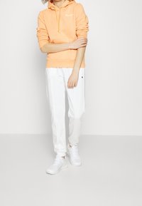 Persikanvärinen huppari, jossa etutasku, valkoisten collegehousujen ja valkoisten lenkkareiden kanssa. Kangas näyttää pehmeältä ja rentoilulta.