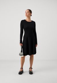 Black long-sleeve dress with a fitted bodice and flared skirt. Features lace accents at the sleeves. Accompanied by a beaded bag and chunky shoes.
