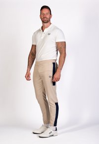 Beige joggers met marineblauwe zijpanelen, elastische tailleband en een voorzak. Gecombineerd met een wit polo-shirt met een logo op de borst.