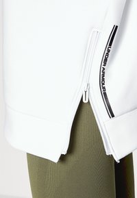 Bílá zipová bunda s černým logem Under Armour, s hladkou texturou a mírně zkrácenou délkou, doplněná olivově zelenými texturovanými legínami.