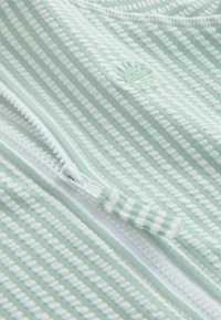 Tela de rayas verde claro con acentos blancos, que presenta una cremallera y un logo de concha en relieve. Textura suave y detalles de patrón acanalado.