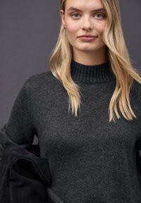 Grå striktrøje med høj ribbet krave, der har en blød tekstur. Lange ærmer, minimalistisk design, parret med et sort plagg.