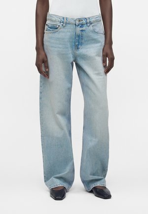 Personne portant un jean bleu clair à jambes larges, un haut blanc sans manches, et des chaussures noires à bout carré pointu sur fond blanc uni.