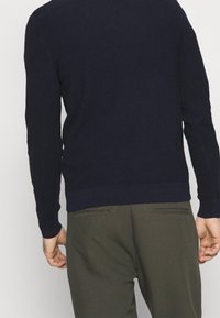 Homme vu de dos portant un pull marine foncé à côtes et un pantalon vert olive avec des poches fendues à l'arrière, debout devant un fond clair uni.