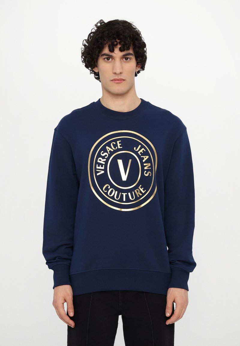 Tamsiai mėlyna džemperis, pagamintas iš medvilnės mišinio, su dideliu auksiniu logo apskritimu, kuriame užrašas "Versace Jeans Couture", ir dubenėlį primenančiais rankogaliais.