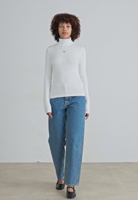 Witte geribde langwerpige turtleneck top gecombineerd met lichtblauwe rechte jeans en zwarte platte schoenen. Minimalistisch ontwerp met subtiele logodetails.