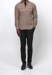 Lichtbruin overhemd met knopen en twee borstzakken, gecombineerd met een zwarte broek en zwarte sneakers. Het overhemd heeft een zachte textuur.