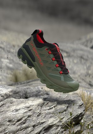 Grüner und schwarzer Trailrunning-Schuh mit roten Akzenten, der über felsigem Gelände mit kleinen Pflanzen und Blumen schwebt.