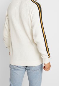 Kremowy sweter z dzianiny z prążkowanymi mankietami i dołem oraz poziomymi kolorowymi paskami w zielonym, pomarańczowym i granatowym kolorze na rękawach.