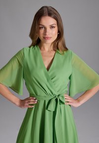 Robe portefeuille verte avec des manches flottantes transparentes, une taille nouée et un tissu fluide. Texture lisse avec un drapé doux, convenant à diverses occasions.