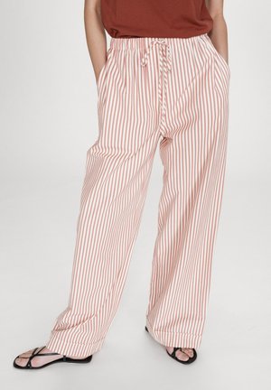 Femme portant un pantalon ample blanc à rayures rouges verticales, taille à cordon, mains dans les poches, et sandales noires à lanières.