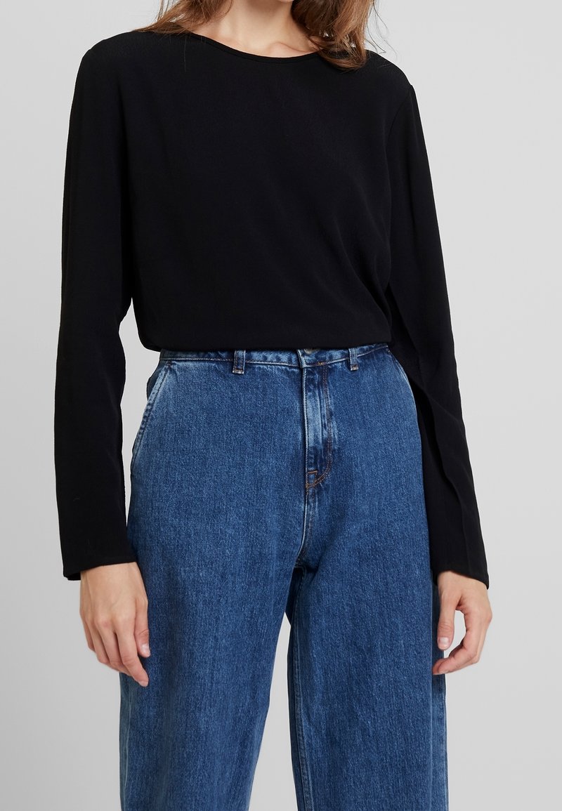 Femme portant une chemise noire à manches longues rentrée dans un jean bleu taille haute avec poches avant.