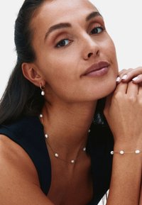 Guldsmycken med pärlor, inklusive ett halsband med flera pärlor, matchande armband och hängande örhängen, visade tillsammans med en mörk topp.